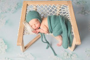 Sleeping newborn baby in teal knit outfit lying on wooden lattice backdrop in professional photo studio setting.