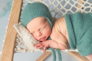 Sleeping newborn baby wearing teal knit wrap lying on white lace blanket during professional photography session.