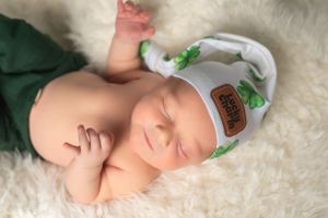 A peaceful sleeping newborn wearing a white and green floral hat rests on a soft white furry blanket.