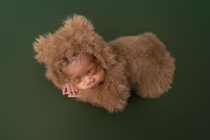 Sleeping baby dressed in a fluffy brown teddy bear costume lies on a dark green background.