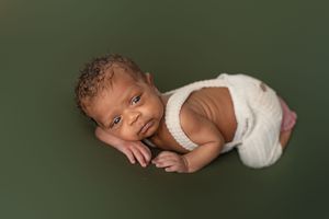 A newborn baby wearing a white onesie lies on a dark green background during a professional photo session.