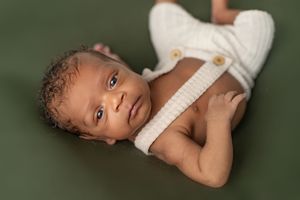 A baby in white clothing lies on a dark green background looking at the camera.