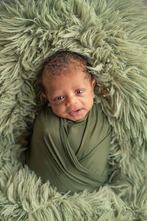 A baby wrapped in olive green fabric nestled in fluffy sage-colored faux fur bedding.