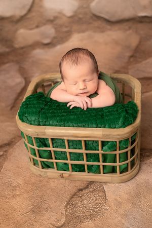 Sleeping newborn rests peacefully on emerald green cushioning inside rustic wooden basket on textured beige backdrop.