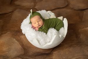Newborn baby sleeping peacefully in white bowl wearing green swaddle on brown backdrop.