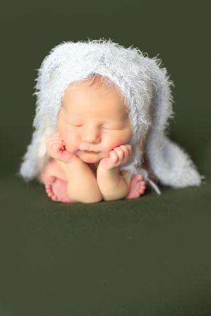 A sleeping newborn baby wearing a white knit bonnet rests with hands tucked under chin against dark green background.