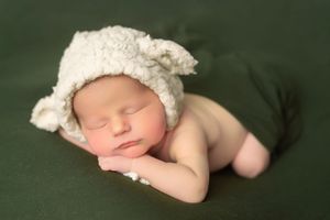 A sleeping newborn baby wearing a white bonnet rests against a dark green backdrop.