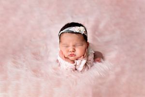 Sleeping newborn wearing a delicate headband rests on soft pink fur backdrop during professional photo session.
