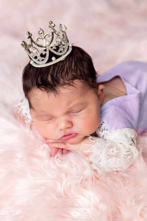 Sleeping newborn wearing silver crown rests peacefully on soft pink fur blanket with white lace details.