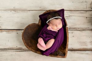 Newborn baby wrapped in purple fabric sleeping peacefully in rustic heart-shaped wooden bowl.