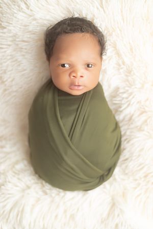 Newborn baby wrapped in olive green swaddle blanket lying on white furry backdrop.