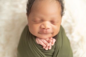 A newborn baby wearing an olive green wrap sleeps peacefully against a cream-colored backdrop.