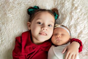 Two children wearing red outfits lie together on white textured fabric, one with green hair bows and one with a headband.