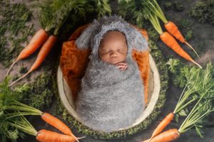 Sleeping newborn nestled in basket surrounded by fresh carrots and greenery.