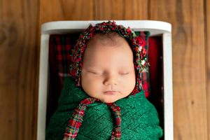 A baby wearing a red and black plaid bonnet and green knit sweater sleeps peacefully against a wooden backdrop.
