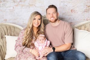 A couple sits on an elegant vintage sofa with their baby wearing pink, against a textured light wall.