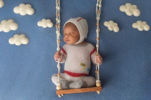 A baby in white clothing swings on a wooden seat against a blue backdrop with cloud decorations.