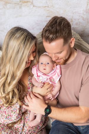 A couple in casual clothing gently holding their baby wearing a pink ruffled outfit against a textured wall background.