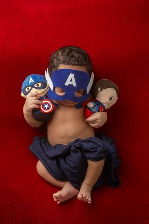 A newborn baby wearing a Captain America themed costume and mask lies on a red backdrop during a photo session.