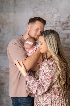 A tender family moment shows caring parents holding their sleeping newborn baby in front of a textured wall background.