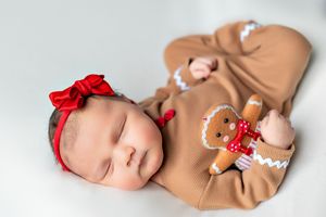 Sleeping baby wearing tan gingerbread outfit with red bow headband and matching plush gingerbread toy.
