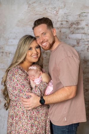 A family poses together against a textured wall background, featuring a floral dress and pink shirt.