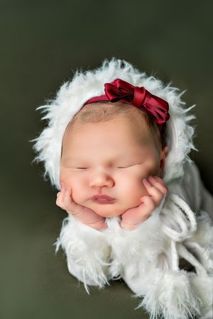 Sleeping newborn wrapped in soft white fur with a red bow headband against dark green background.