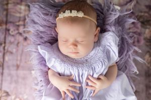 Sleeping baby wearing delicate purple smocked dress and pearl headband on floral backdrop.