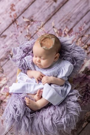 Sweet baby gently resting on soft purple faux fur with delicate lace crown and white dress, photographed from above.