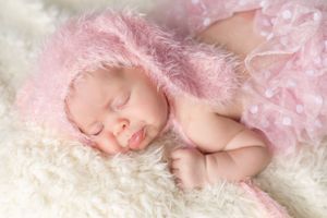 Sleeping newborn wearing soft pink feathery headband and tutu lying on fluffy white blanket in dreamy lighting.