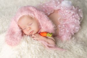Sleeping newborn wrapped in soft pink lace lying on white fluffy blanket with small orange plush toy.