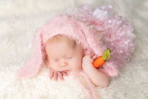 Peaceful sleeping newborn in soft pink blanket and headpiece lying on white furry backdrop holding small carrot toy.