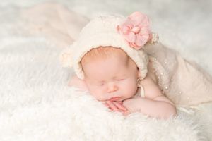 A sleeping newborn wearing a cream bonnet with pink flower rests peacefully on soft white bedding.