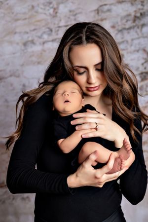 Tender moment between mother and sleeping newborn in black against brick wall.