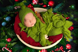 A newborn in bright green knit outfit sleeps in a white bowl surrounded by Christmas decorations and candy canes.