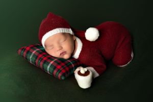 Sleeping newborn in red Santa outfit on plaid pillow against dark green background.
