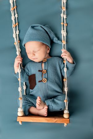 Sleeping newborn in blue knit outfit rests peacefully on decorative wooden rope swing against teal backdrop.