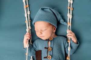Sleeping newborn wearing blue knit outfit and hat rests peacefully on macrame swing against teal backdrop.