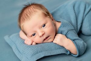 Newborn baby wearing soft blue knit sweater lying on blue blanket while resting head on hand.