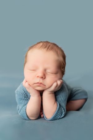 Sleeping newborn in light blue outfit rests peacefully with hands under chin on blue backdrop.