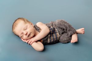Sleeping baby wearing plaid overalls lies peacefully on blue backdrop during professional newborn photoshoot.