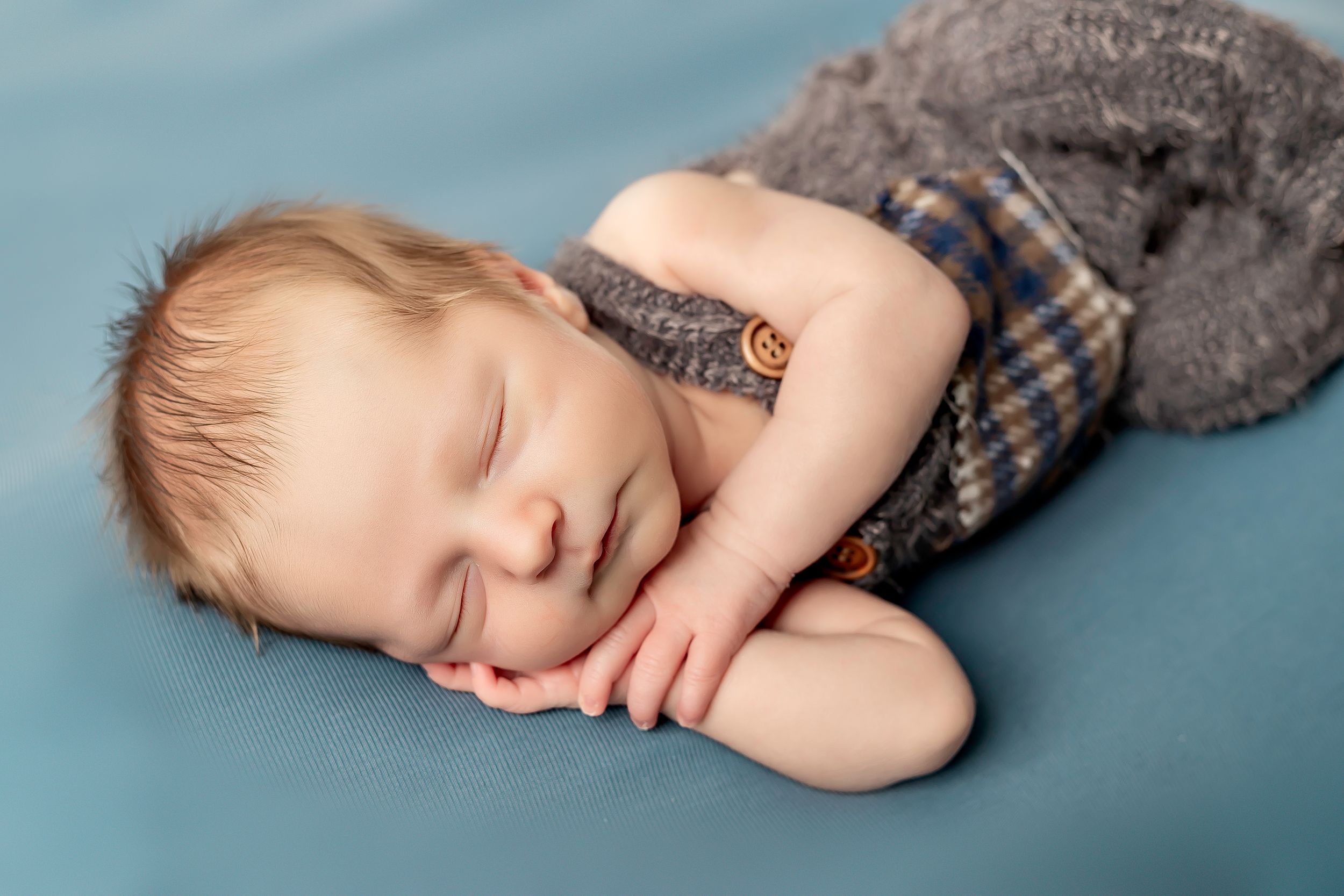 A sleeping baby in a gray sweater and plaid shorts rests peacefully on a teal backdrop.