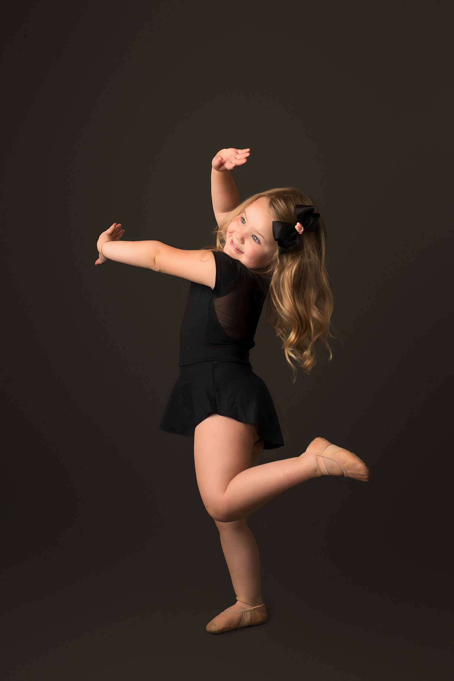 Young dancer in black leotard strikes graceful pose against dark background.