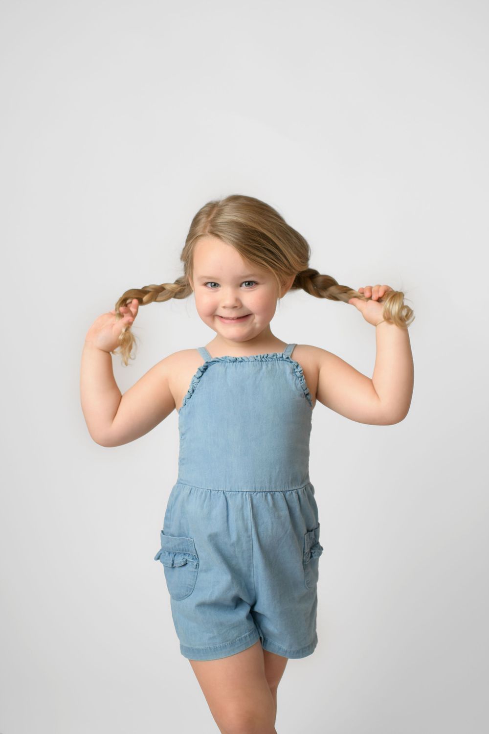 Young child in blue romper playfully pulls pigtails while smiling at camera.