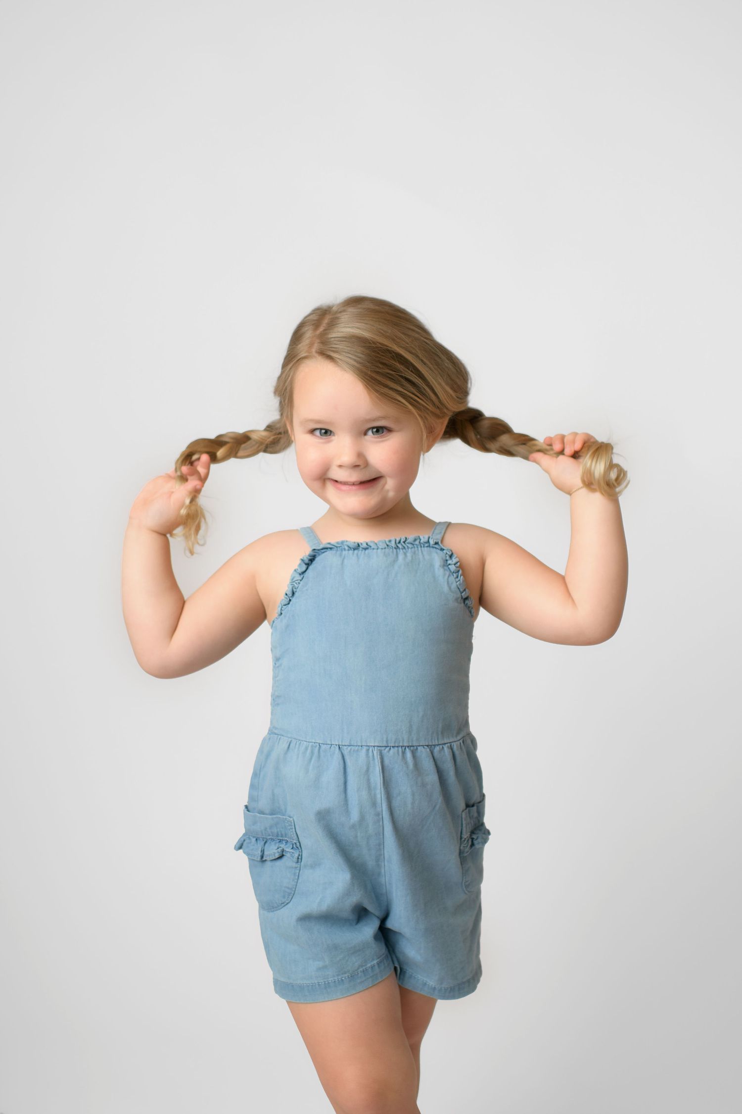 Young child in blue romper playfully pulls pigtails while smiling at camera.