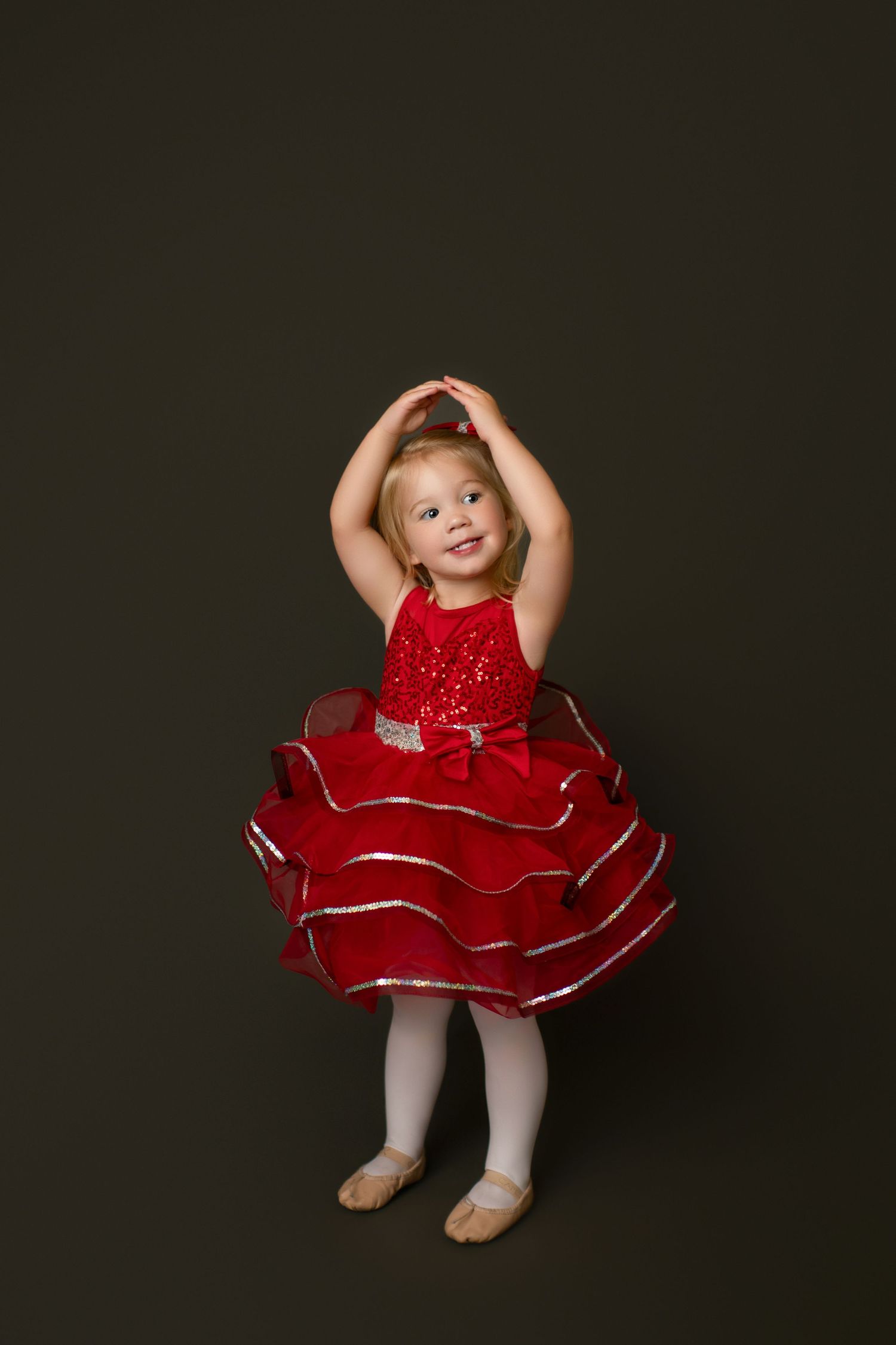 Young dancer in sparkly red dress poses with arms raised against dark background.