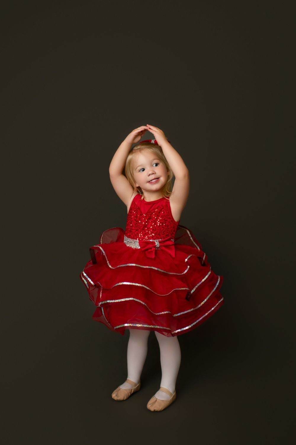 Young dancer in sparkly red dress poses with arms raised against dark background.