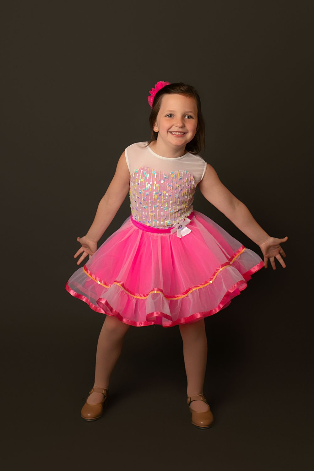 Dancer poses in pink tutu dress with sequined bodice against dark studio background.