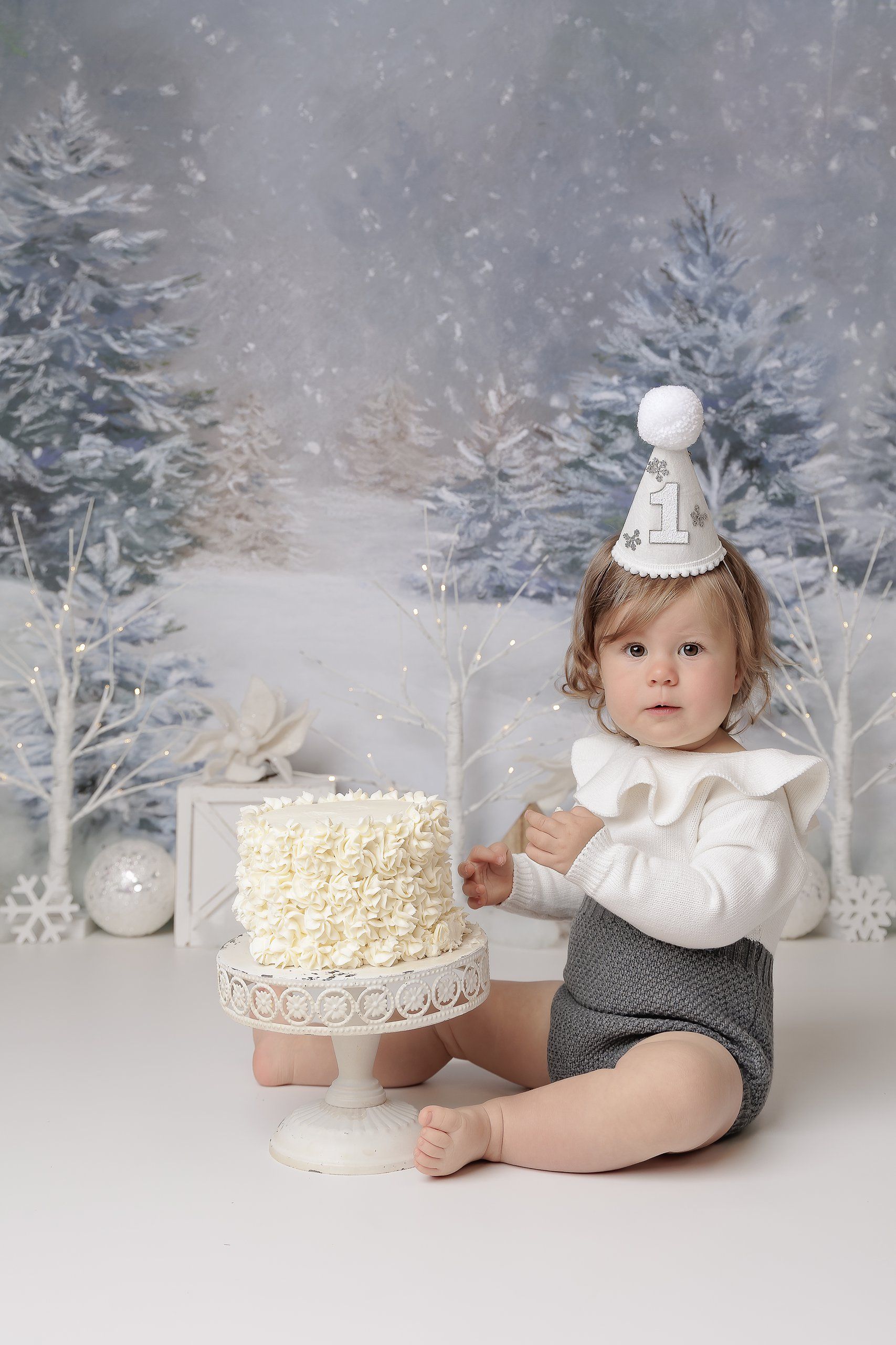 Baby girl in snowy forest set with gray and white knit outfit with ruffle collar wearing a snowflake embellished birthday hat and cake.