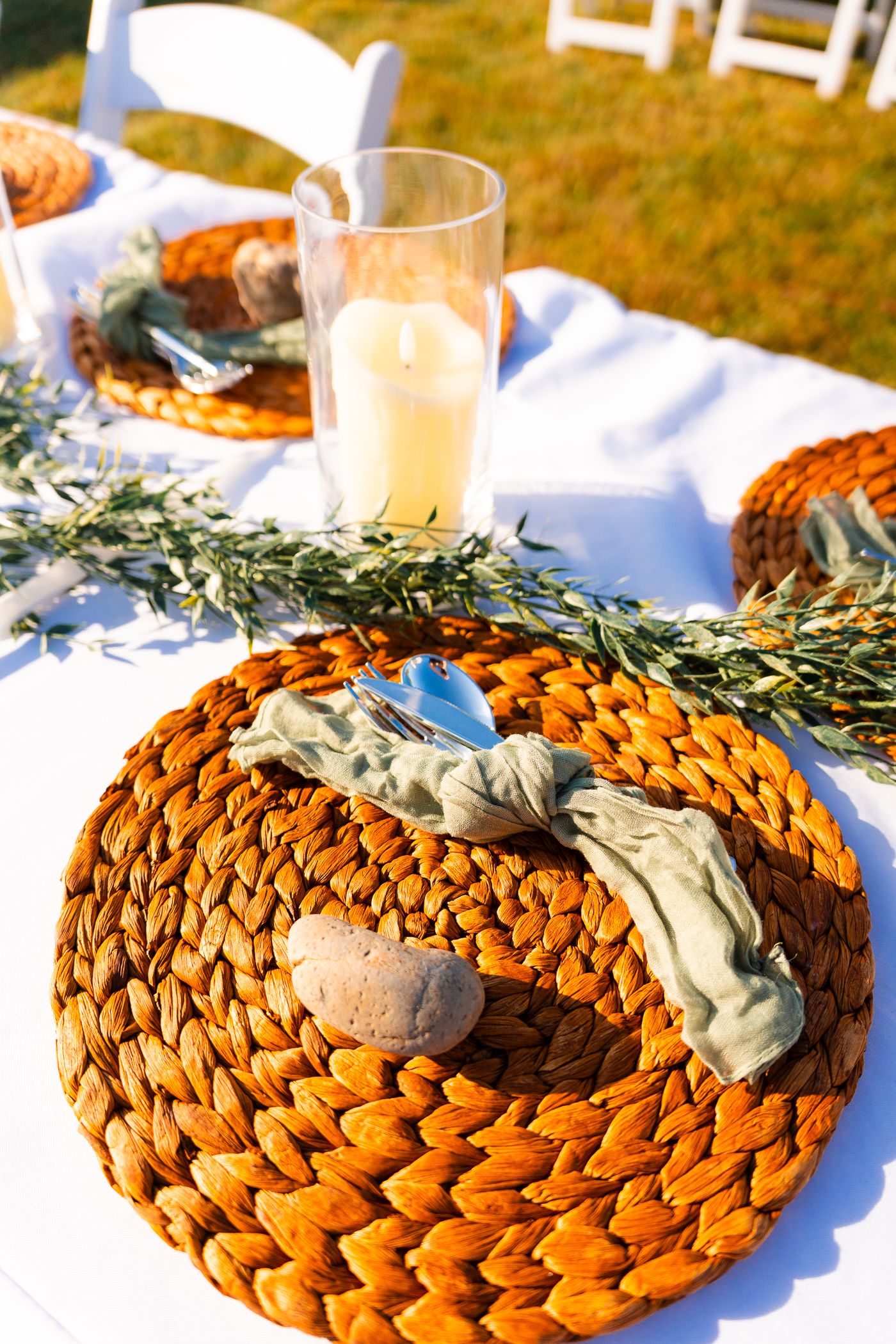 Woven rattan charger plate with eucalyptus sprig and napkin on white tablecloth with glass in background.